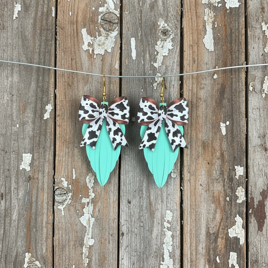 Roselee Glitz mint faux leather leaf earrings with cow print fabric bows and gold hardware on rustic wood background