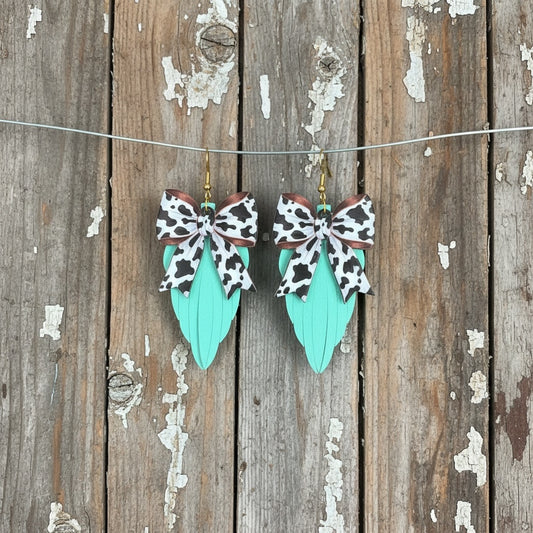 Roselee Glitz mint faux leather leaf earrings with cow print fabric bows and gold hardware on rustic wood background